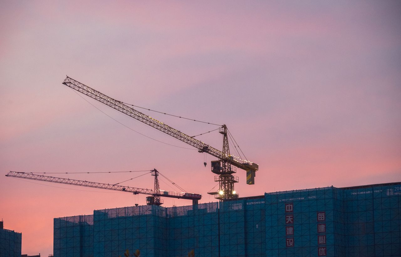 Construction cranes tower over a city skyline at sunset, symbolizing urban growth.